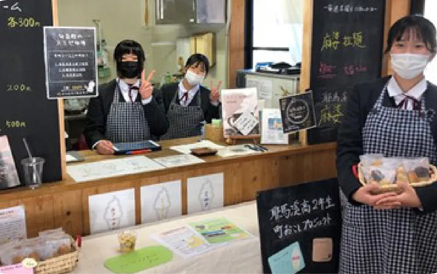 高校生カフェのレジカウンターに立つ3人の女子学生。一人はクッキーの箱を持っており、他の二人はピースサインをしている。