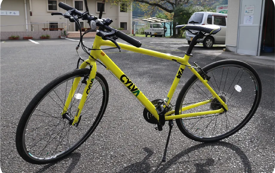 黄色い自転車が屋外に置かれている。背景には、建物や軽トラックが写っている。