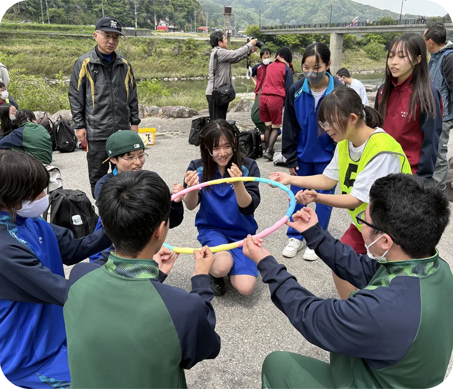 屋外でフラフープを使って輪を作っている5人の学生。周囲には他の学生や大人がいる