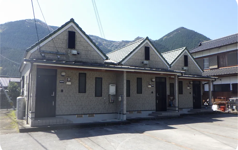 三角屋根が並んだ二階建て風のデザインをした、3戸が連なるアパート。背後には山々が広がっている