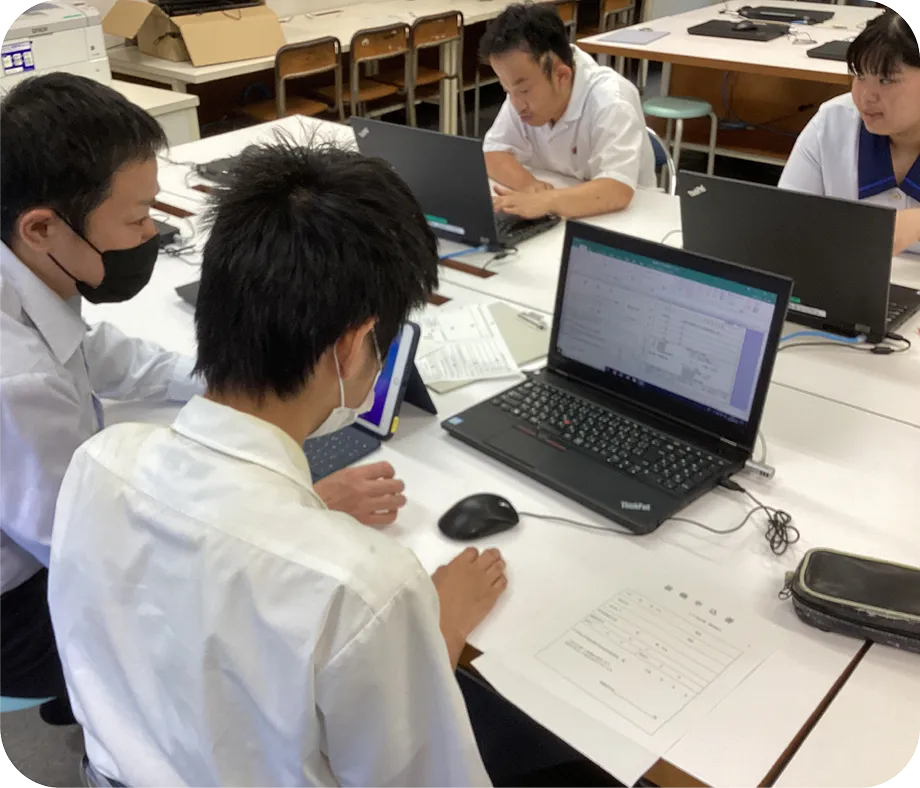  4人の学生と1人の教師が、パソコンが置かれた机を囲んで座っており、教師はパソコンの画面を覗き込みながら、学生にアドバイスをしている。