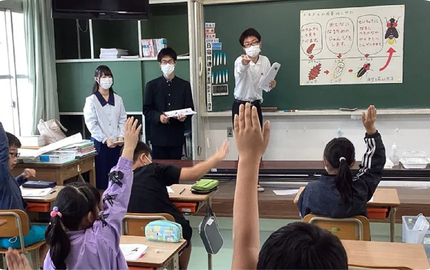 教室で小学生に授業をしている3人の高校生。一人がマイクを持ち、手を挙げている小学生に向けている。