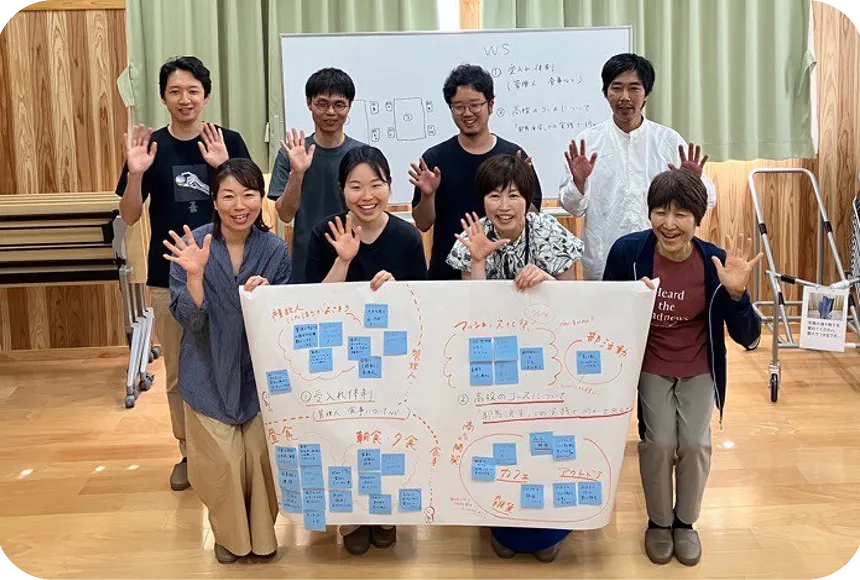  室内に集まった7人の大人と学生が笑顔で手を振っており、そのうちの4人が、青い付箋がたくさん貼られた大きな紙を掲げている。
